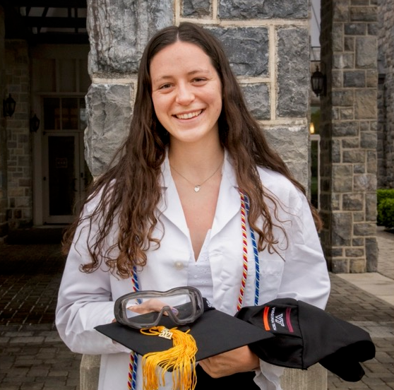 Tess Alexander, B.S. in Biology, with a minor in National Security & Foreign Affairs, is smiling, wearing a lab coat and holding her graduation cap and safety goggles.