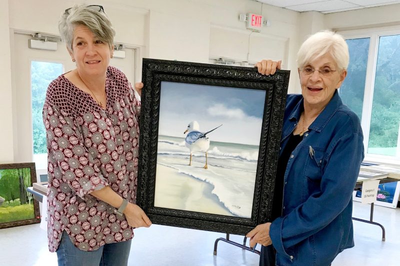Two women hold up a oil painting of a sea gull on the beach.