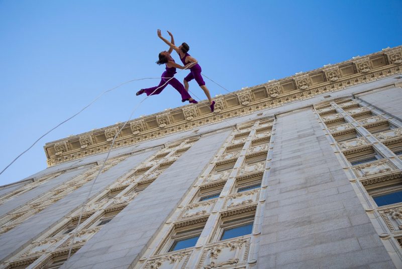 Two dancers in purple costumes hang onto separate ropes and gracefully rappel from the side of an ornate building with lots of windows. The perspective is from the ground, looking up at the length of the building.