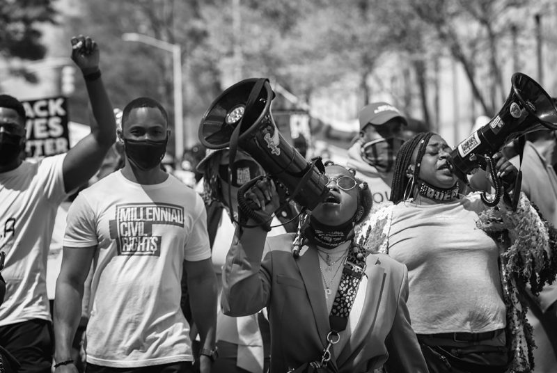 A black and white image by photographer Sheila Pree Bright from a voter suppression rally in Atlanta, Georgia,  shows several protestors marching together down a street, some with megaphones.