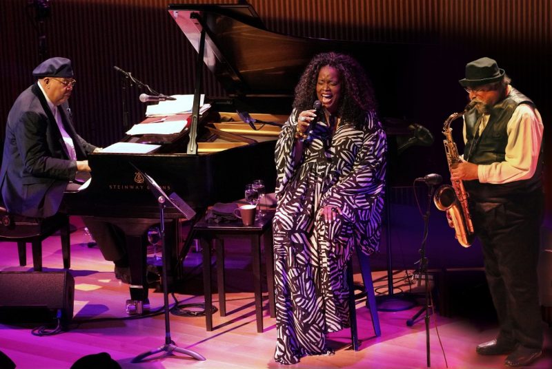 Jazz vocalist Dianne Reeves sits at the center of a stage sitting on a stool, flanked by Chucho Valdés on the left playing piano and Joe Lovano on the right playing saxophone.