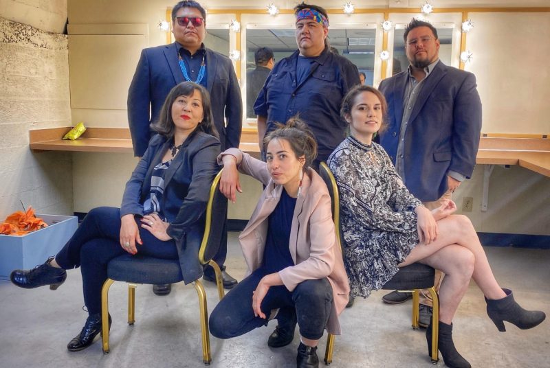 The six artists/musicians that perform for the production "Welcome to Indian Country" pose in a dressing room - three men stand, while two women sit in chairs with a third woman kneeling between the two sitting.