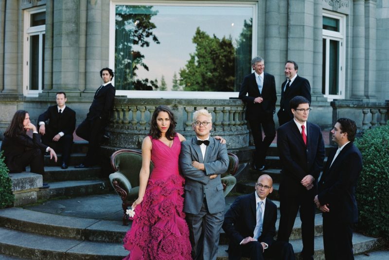Members of the ensemble Pink Martini pose outside on the steps of a stone building. Some members in suites sit on steps and lean on ledges, while a woman in a hot pink, frilly dress poses with her arm around a man in a suit and bowtie.