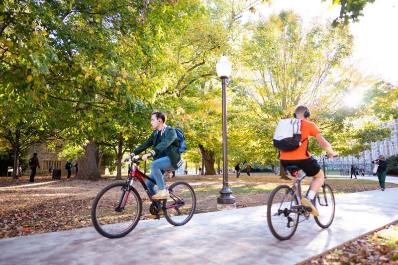 Students ride bikes in opposite directions against a fall foliage backdrop