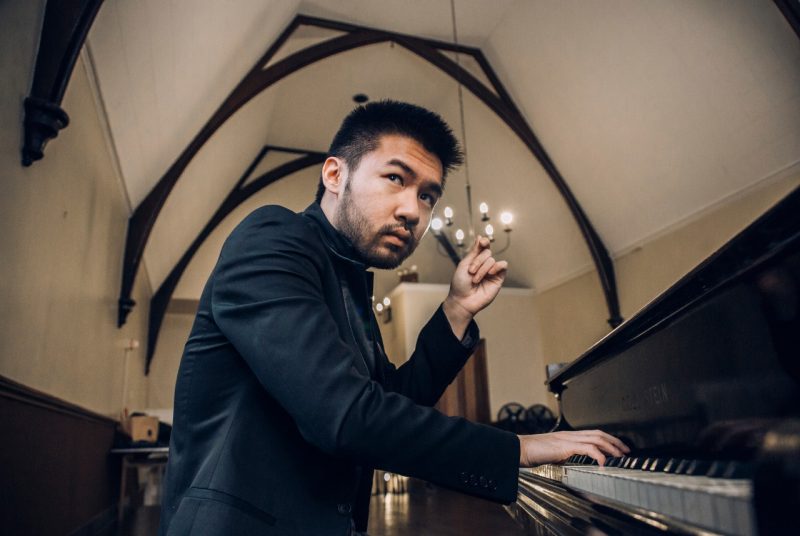 Conrad Tao, an Asian man with dark hair and a closely trimmed beard, sits at a piano in a dark blazer.