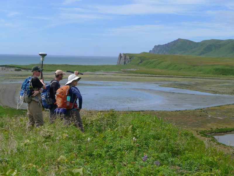 Three people standing on a grassy hill, overlooking marshland and the Alaskan coast.