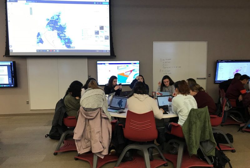 A group of ten students sits around a table, talking and working on laptop computers, with several large screens on the wall behind them