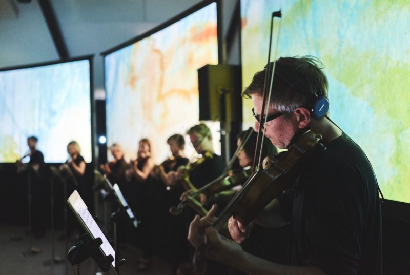 Musicians with Scottish Ensemble are lined up in a row, watching sheet music on stands in front of them while standing in front of three large screens showing abstract scenes.