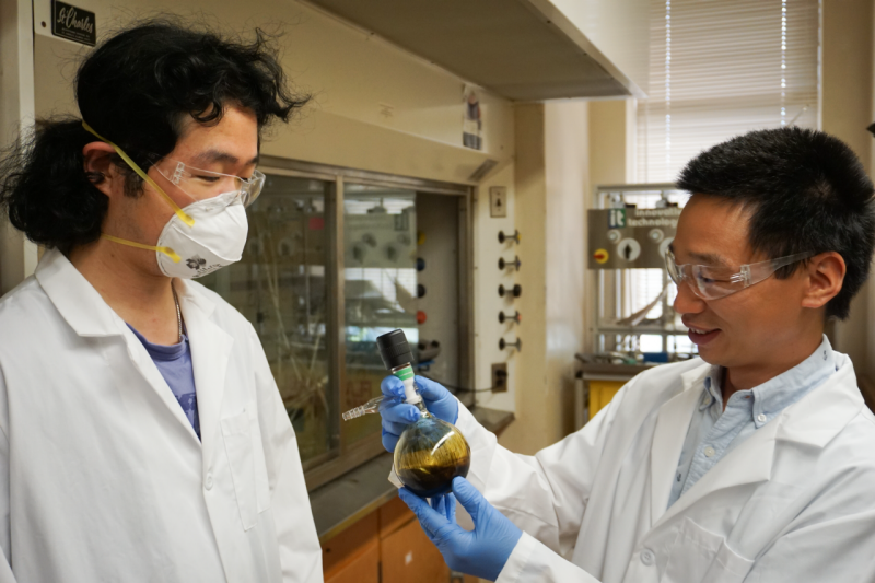 Two men wearing lab coats smile while looking at a small glass container.