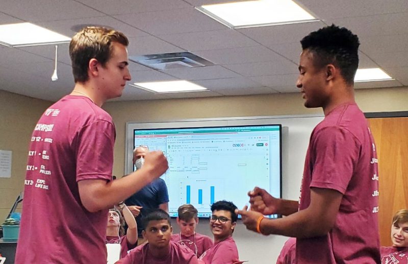 Two standing male students, surrounded by sitting students, play a game of Rock, Paper, Scissors.