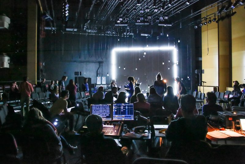 A view from the side of a stage shows actors and technicians sitting in chairs and looking at computer screens, rehearsing for a production. They are all backlit by various lights hanging from the ceiling and a glowing square against a black wall.