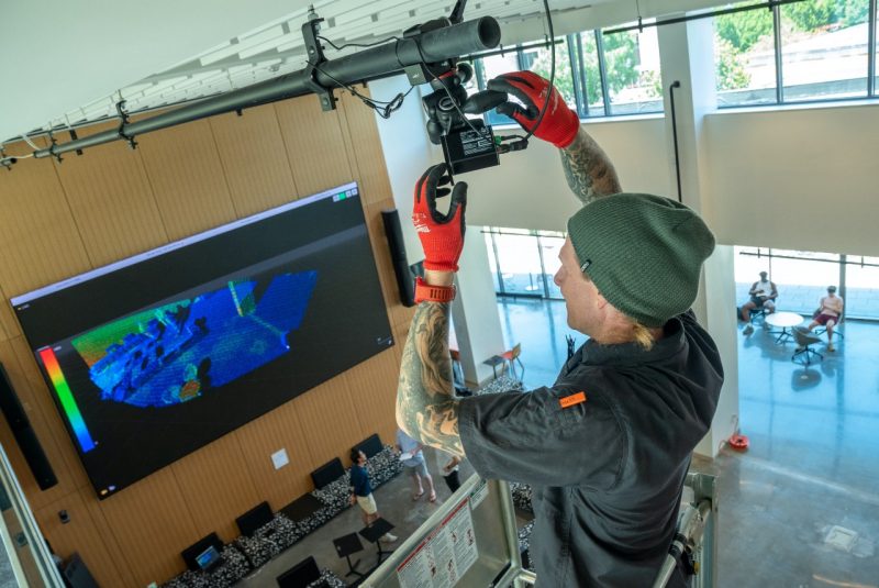 A man installs a lidar sensor in the ceiling of the Creativity and Innovation District, with the lobby area in the background. 
