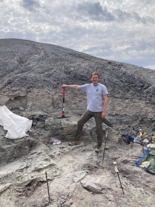 A man  wearing  a t-shirt and jeans holds the handle of a pick ax. Behind him is the hard ground of the Petrified National Forest.