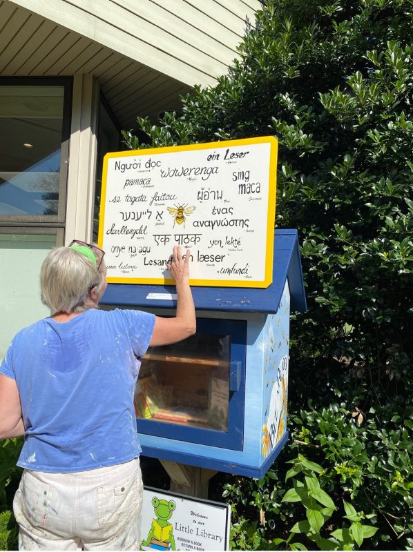 A woman stands in front of a colorful box filled with books with her back to the camera. She holds a hand up to a sign in multiple languages reading "little free library."
