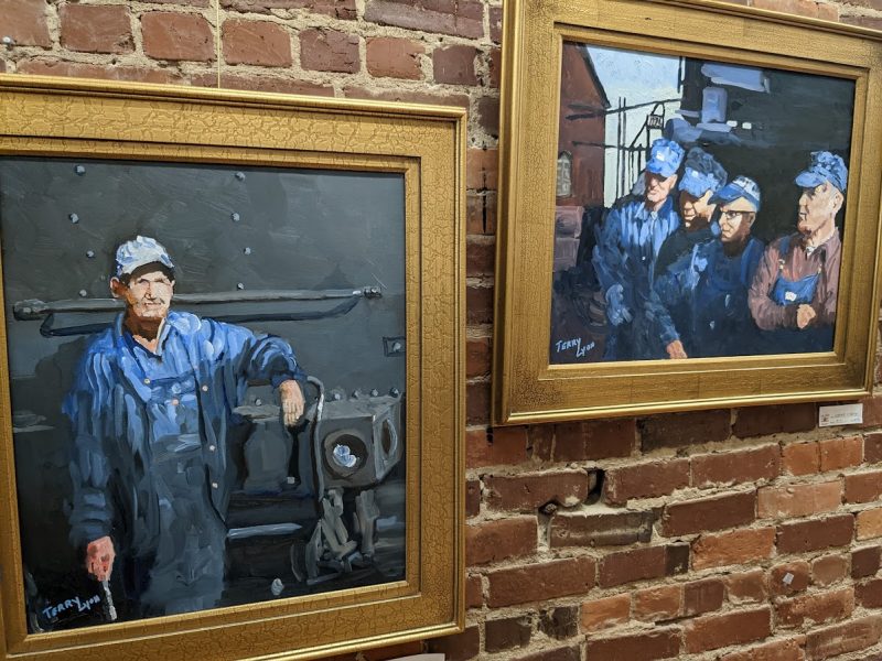 Two paintings with gold-painted frames mounted on a red brick wall. The left one depicts a mechanic in a blue jumpsuit leaning against a black train engine. The right depicts four railworkers in overalls and blue hats posing in front of a roundhouse.