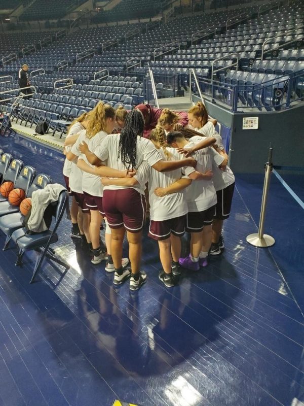 The women’s team huddles together in a circle in the basketball arena. 