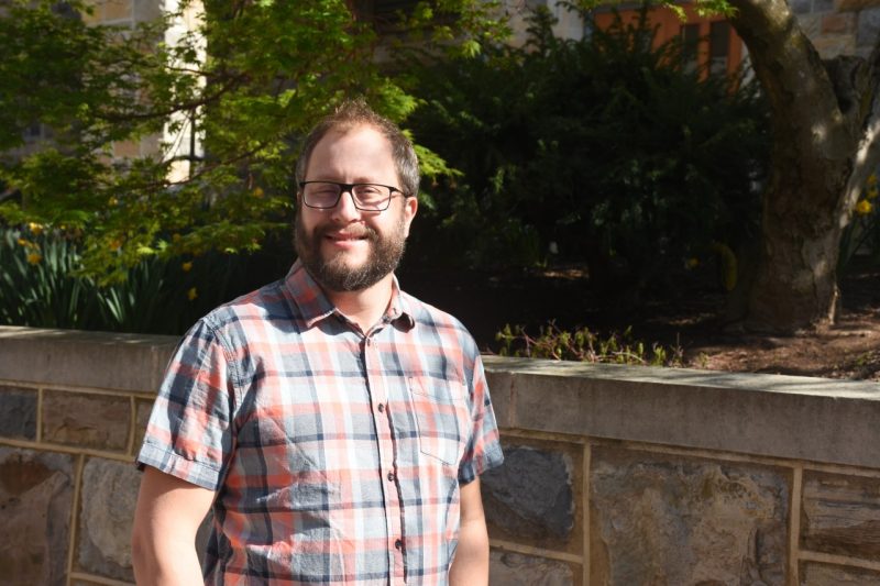 A man with. reddish hair and a beard and glasses, poses in. checkered shirt near shrubbery and Hokie Stone walls.