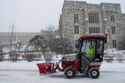 A mini red snow plow removes snow on a roadway in front of grey Hokie Stone Newman Library on a snowy day. 