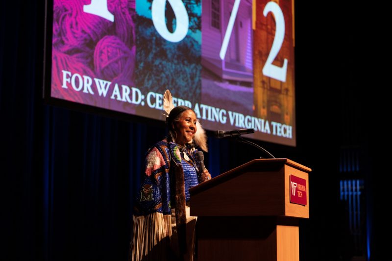 Victoria Ferguson, member of Monacan Nation, appears at a podium wearing traditional Native American attire. She is holding a microphone and stands in front of a colorful background.