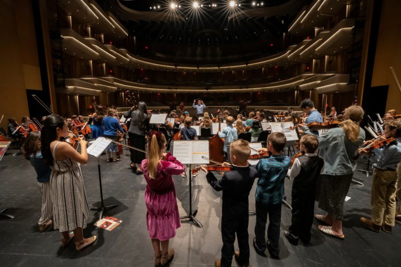 Several rows of children in varying degrees of formal dress play string instruments on the stage of Moss Arts Center with music stands in front of them. Rows of lit balconies are visible in the background, and bright lights are visible at the top of the image.