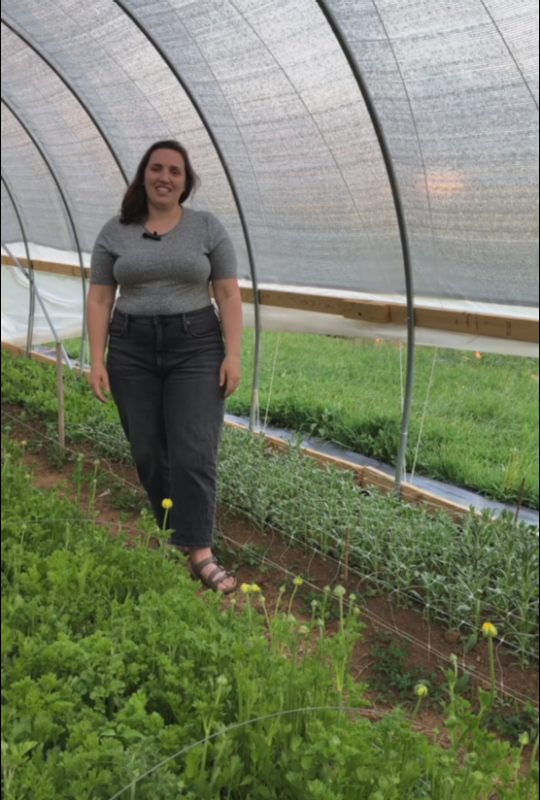Woman in gray shirt stands in a greenhouse lined with plants.