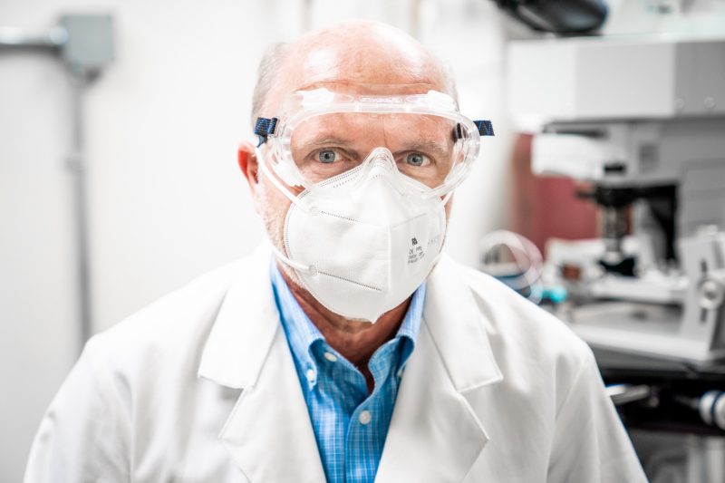 man looks at camera in laboratory wearing goggles, face mask, and lab coat